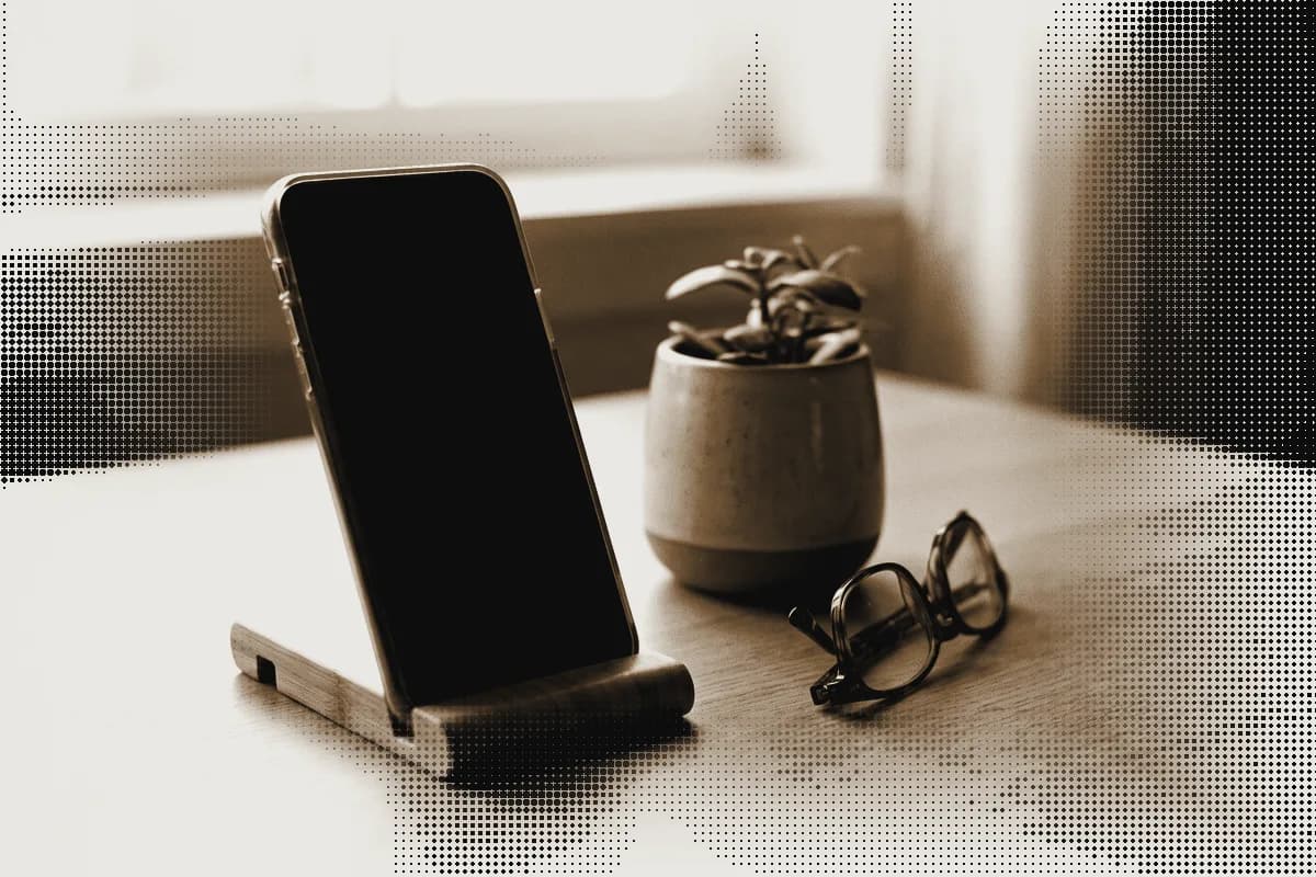 A phone standing in a wooden stand beside a small plant and a pair of glasses, pressed in bronze
