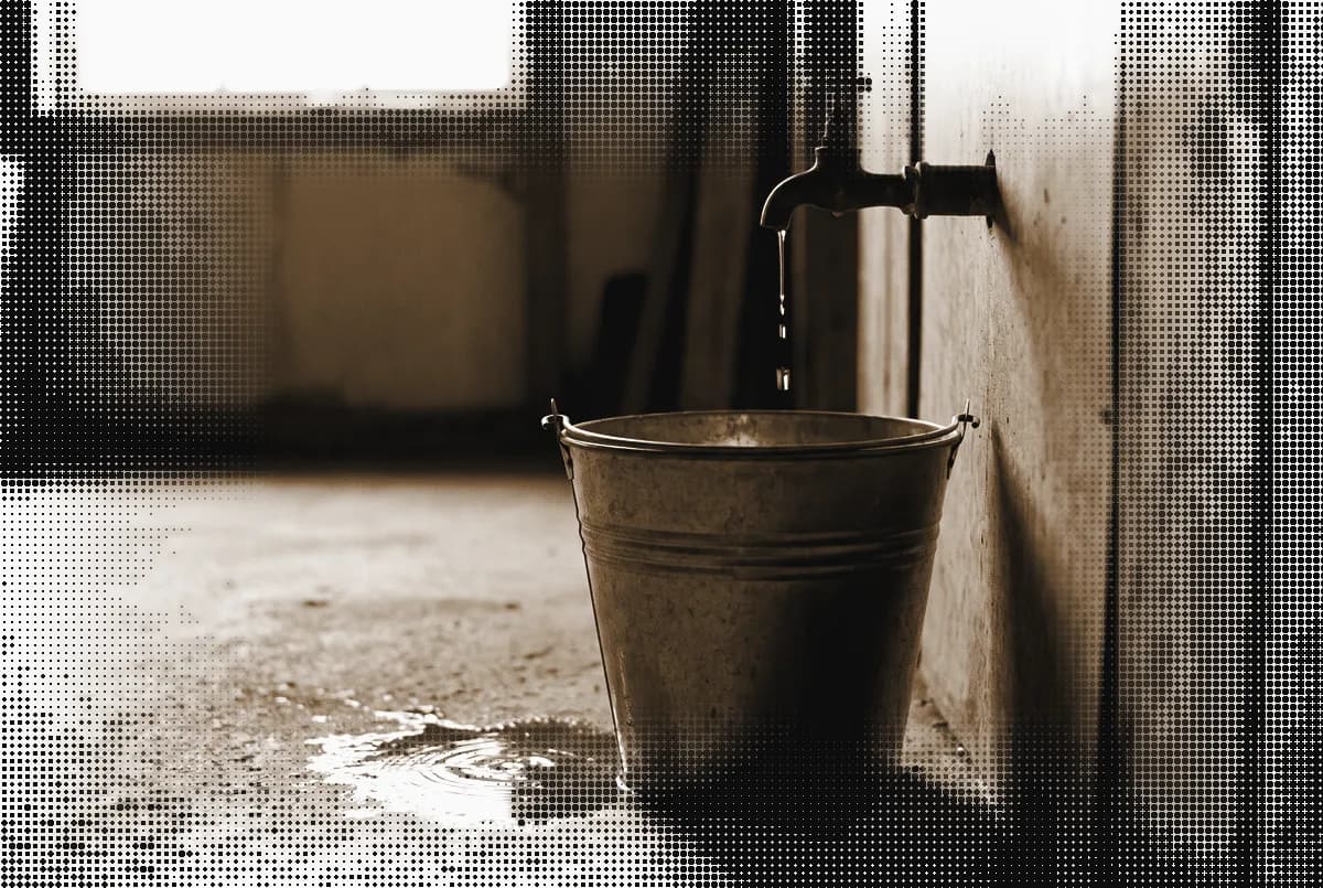 A galvanized bucket under a dripping brass tap on a workshop floor, pressed in bronze