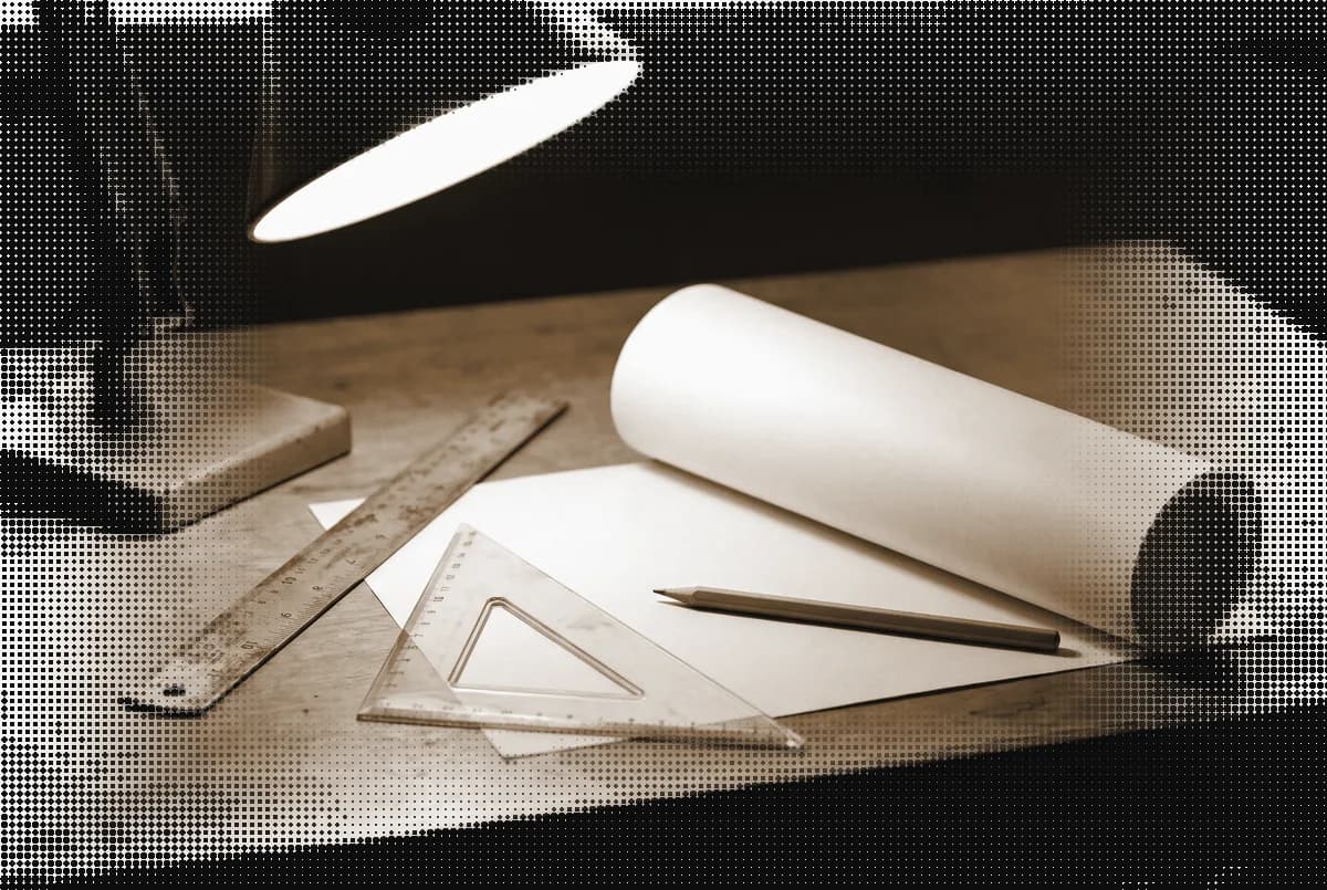 A drafting table with a steel ruler, a set square, a rolled sheet and a pencil under a lamp, pressed in bronze
